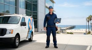 Professional commercial electrician standing in front of a modern building in Hermosa Beach, California, with ocean and pier in background, holding a tablet